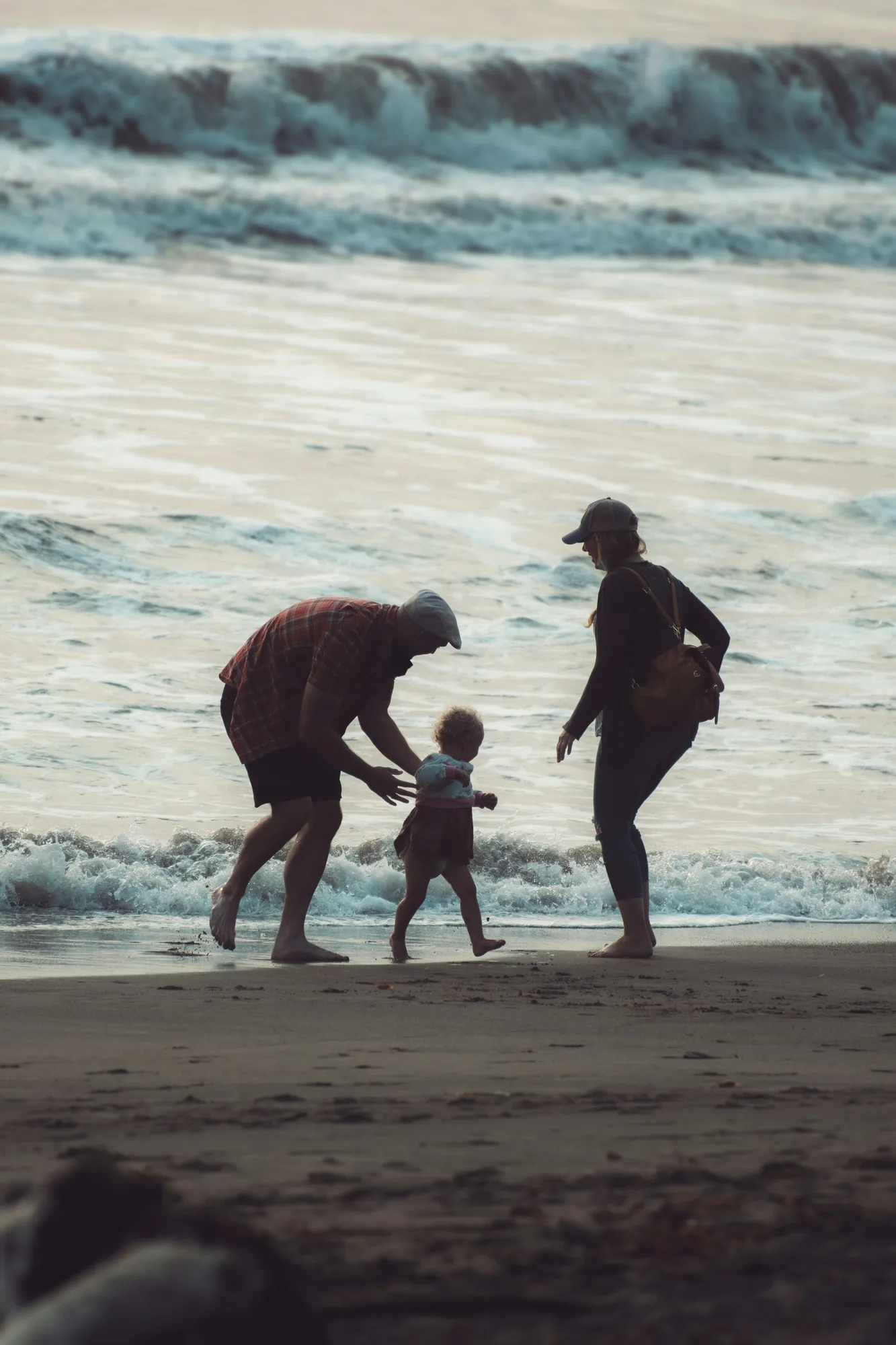 A family helping a toddler take steps at the water’s edge