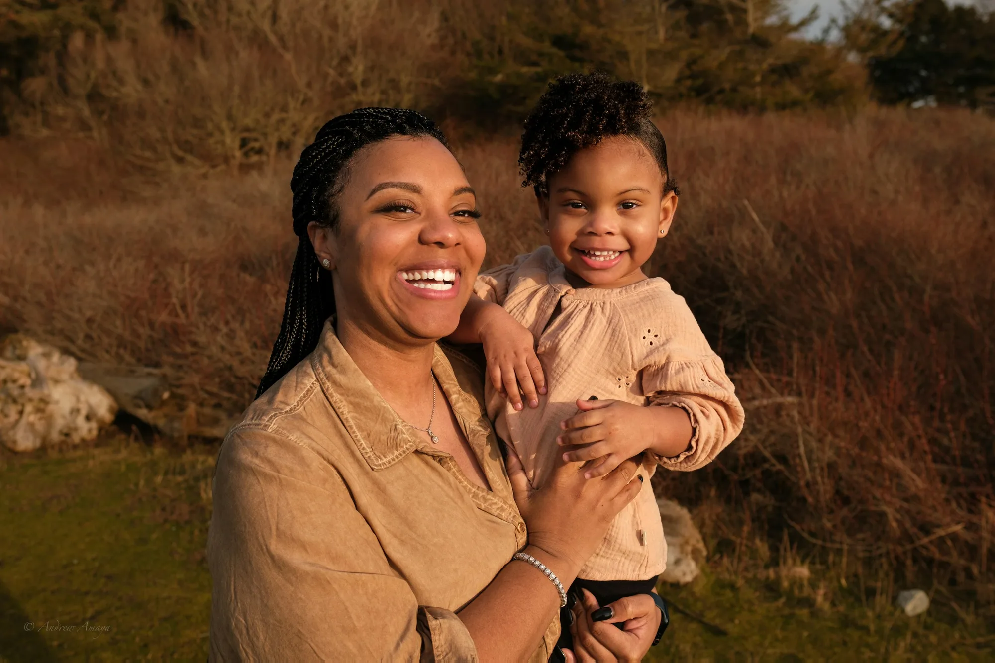 A parent smiling with their young daughter in her arms, outdoors at golden hour