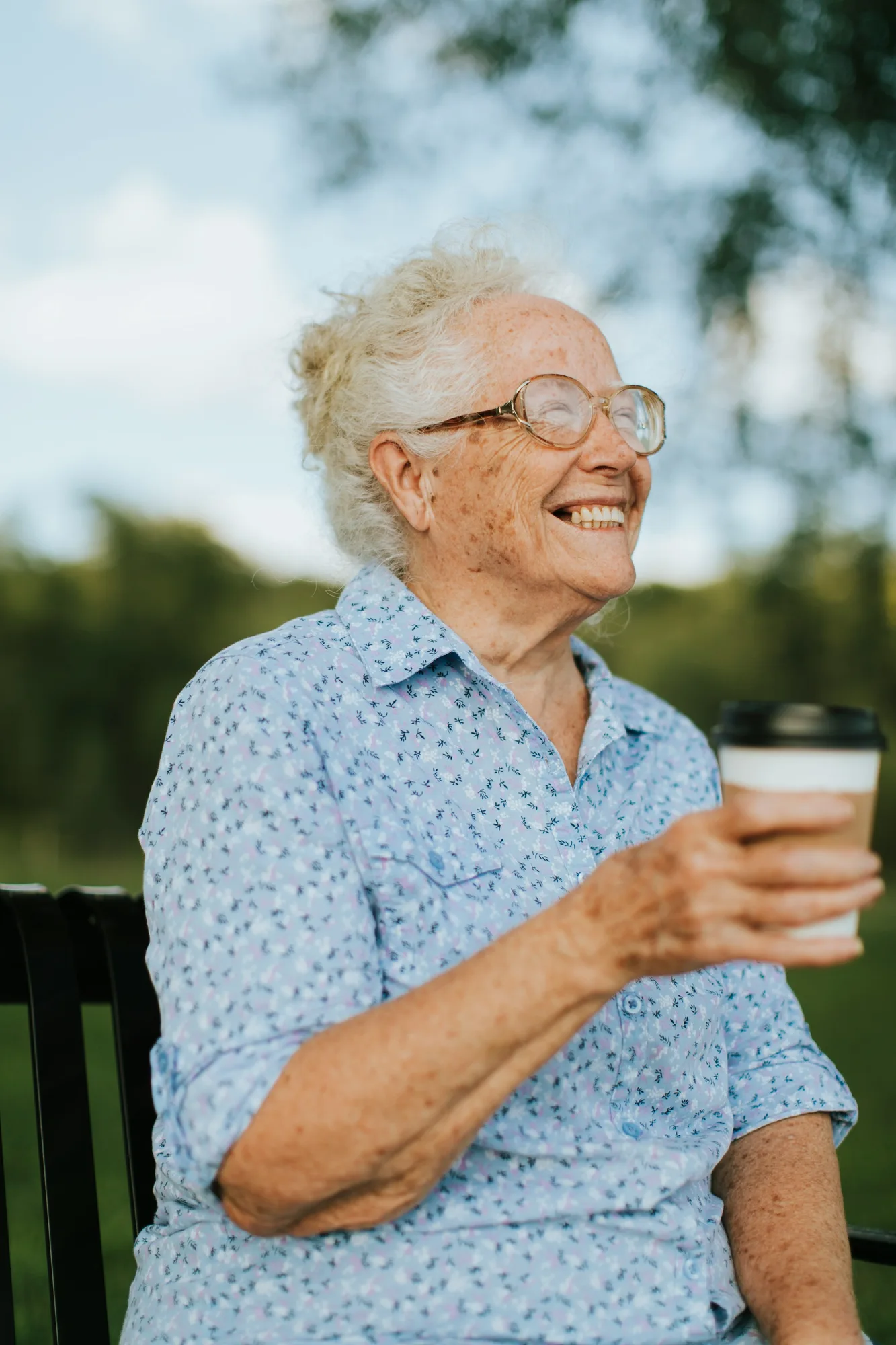 An older woman laughing on a park bench, holding a cup of coffee